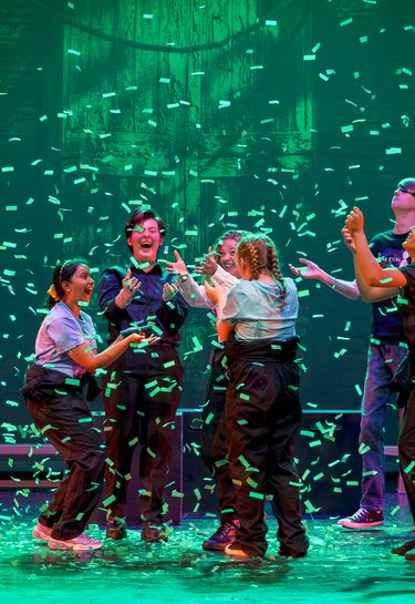 Six young performers on stage are light by a emerald green light as confetti falls to the ground and the jump in exhilaration. This photo is from the 2023 Cast Youth Theatre production of Time Casters.