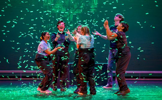 Six young performers on stage are light by a emerald green light as confetti falls to the ground and the jump in exhilaration. This photo is from the 2023 Cast Youth Theatre production of Time Casters.