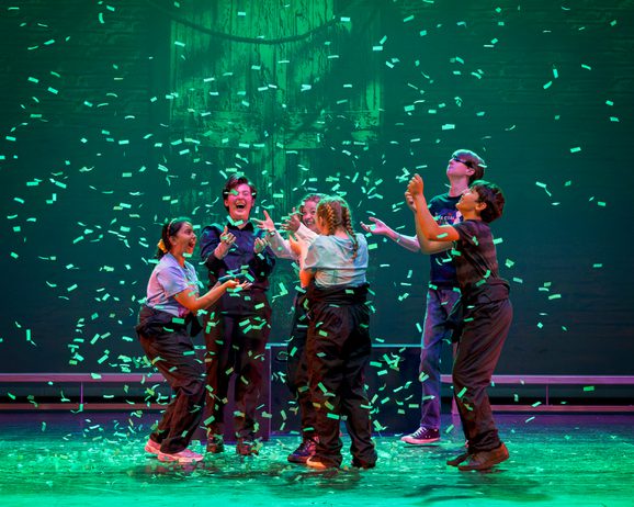 Six young performers on stage are light by a emerald green light as confetti falls to the ground and the jump in exhilaration. This photo is from the 2023 Cast Youth Theatre production of Time Casters.
