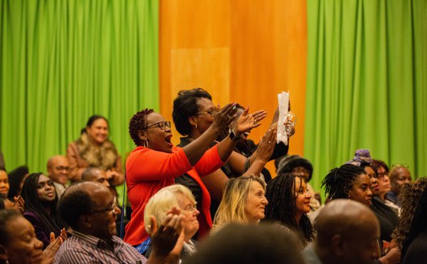 Audience members, predominantly people of color, applaud enthusiastically in a brightly lit room with green and orange drapes.