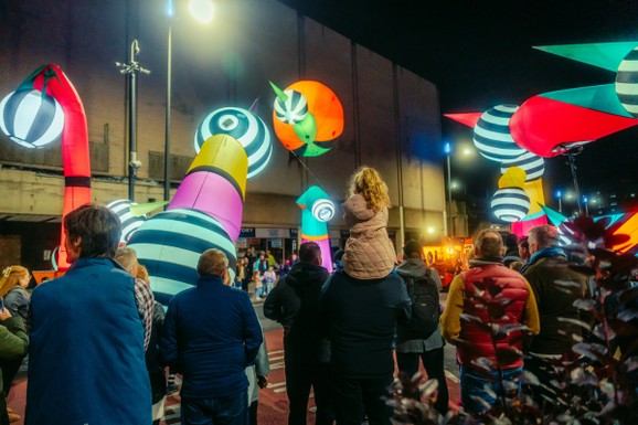 Crowd gathers around colorful, illuminated, inflatable giants which are characters in a piece of promenade theatre at a night-time event.