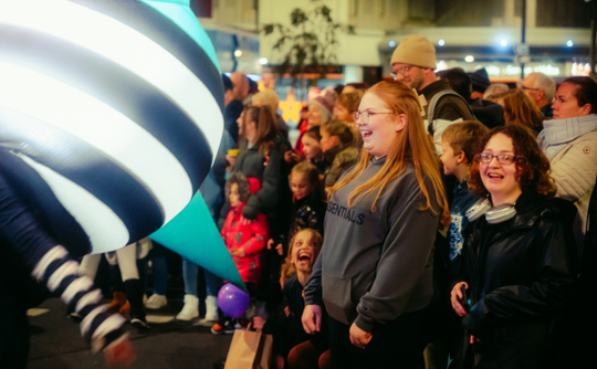 A joyful crowd at a night event watches an oversized, striped balloon pass by. Faces are lit with amusement and wonder, creating a festive atmosphere as the audience watch Lampadophores. A Global Streets performance.