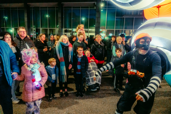 A diverse group of people, including children, are joyfully watching a performer in a striped costume and orange hat during a night outdoor performance of Lampadophores at Global Streets..