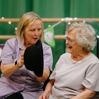 An elderly woman with gray hair and glasses laughs as a caregiver in a lavender uniform presents her with a black bowler hat. The two women are seated, together, with festive bunting and lights in the background.