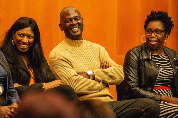 Three people sit in a room with wood paneling, smiling and laughing.