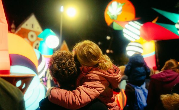 A adult women with curly hair and young child with blond hair are hugging as they look on to a variety of giant balloon like characters from the performance of Lampadophores.