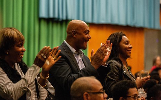 A group of people clapping and smiling in an auditorium with green and blue curtains. The mood is joyful and supportive.