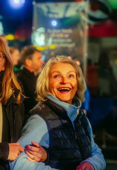 A joyful group of three people stands in an illuminated crowd at night, smiling and linking arms. The atmosphere is warm and joyful during the 2024 Global Streets performance of Lampadophores.