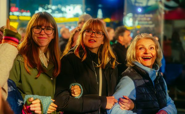 A joyful group of three people stands in an illuminated crowd at night, smiling and linking arms. The atmosphere is warm and joyful during the 2024 Global Streets performance of Lampadophores.