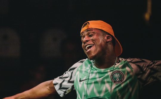 A young Black man in an orange cap and green football t-shirt smiles with arms spread.