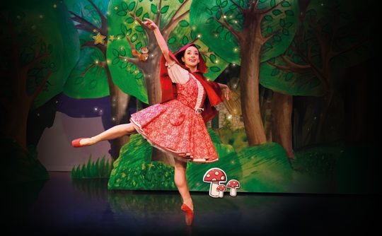 A ballerina in a red outfit joyfully leaps on stage, set against a whimsical forest backdrop with painted trees and mushrooms, conveying a fairy tale vibe.