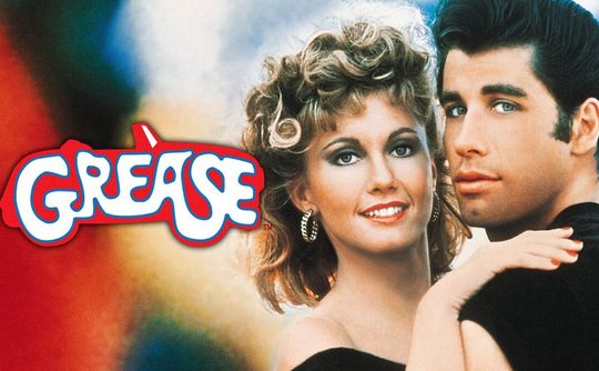 Couple from the movie "Grease" embraces against a colourful background. The logo "Grease" is prominently displayed in bold, stylized white text.