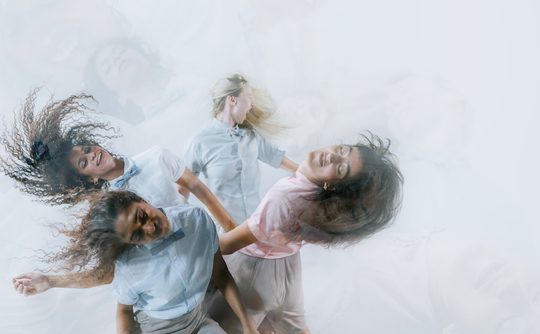 Three women joyfully dance, their hair and pastel shirts swirling in motion. The blurred background and smiles convey energy and happiness.
