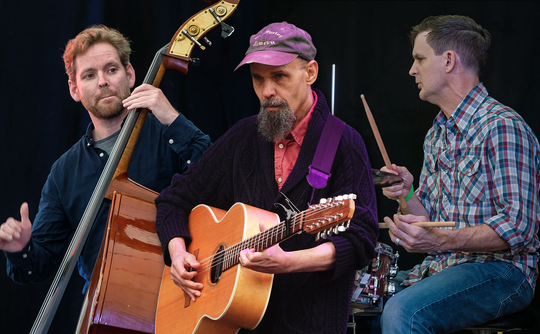 Three musicians perform on stage: one plays a double bass, another strums an acoustic guitar, and the third plays drums. The scene feels lively and engaged.
