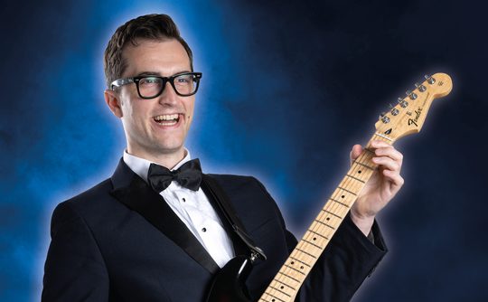 A smiling man in a tuxedo and glasses plays an electric guitar against a blue smoky background, conveying a joyful and lively atmosphere.