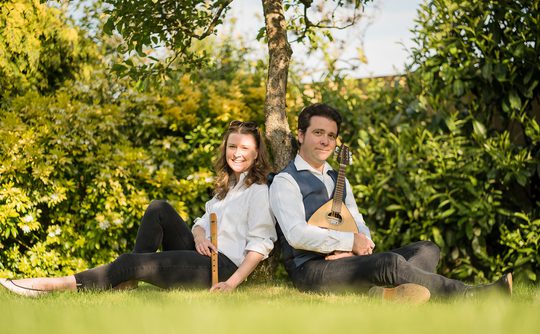 A man and woman sit back-to-back under a tree in a sunny garden. The woman holds a flute and smiles, while the man holds a mandolin, looking content.