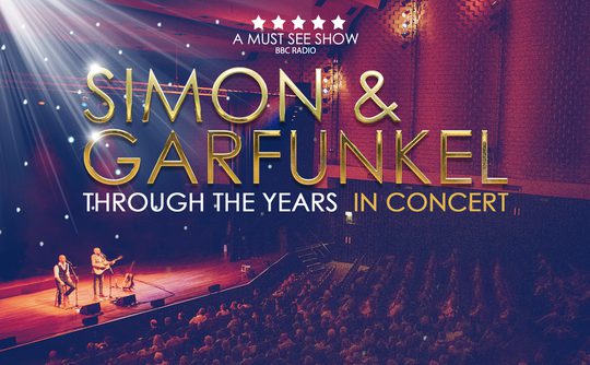 Concert image with two musicians onstage under spotlights, facing a large audience. Text overlay reads, 'Simon & Garfunkel Through the Years In Concert.' The setting conveys a nostalgic and lively atmosphere.