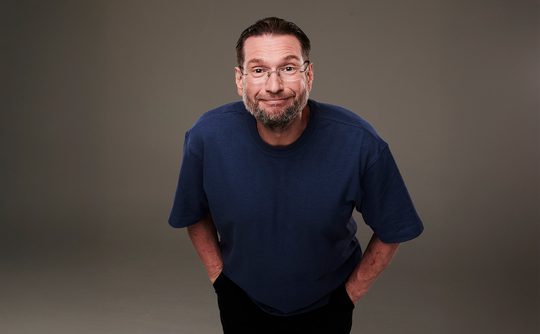 A man with glasses and a beard leans forward, smiling warmly. He wears a blue T-shirt against a plain grey background, conveying a friendly, casual vibe.