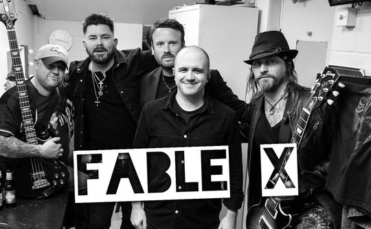 Black and white photo of five men, some holding musical instruments, smiling at the camera. The text "FABLE X" is prominently displayed in front.