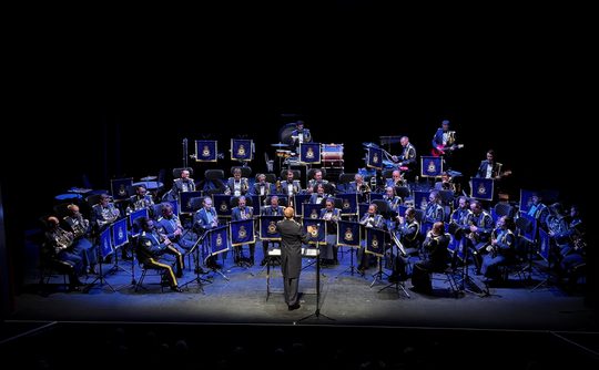 A large orchestra performs on stage, featuring musicians in uniform with blue music stands. A conductor stands in front, creating a formal and focused ambiance.