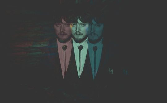 A man in a suit is depicted in a triple-exposure effect against a dark background, creating an eerie, surreal aesthetic with varying shades and emotions.