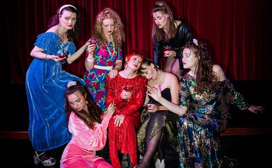 Seven women in colourful, vintage dresses pose dramatically against a red curtain. They each hold a glass, creating a theatrical, nostalgic vibe.