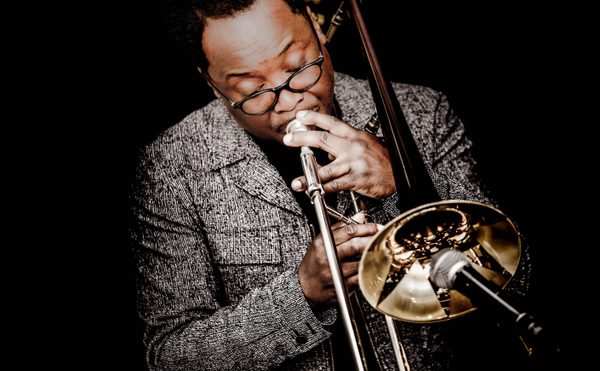 Man playing trombone with focus and passion, wearing glasses and textured jacket against dark background. Microphone in foreground, conveying musical atmosphere.