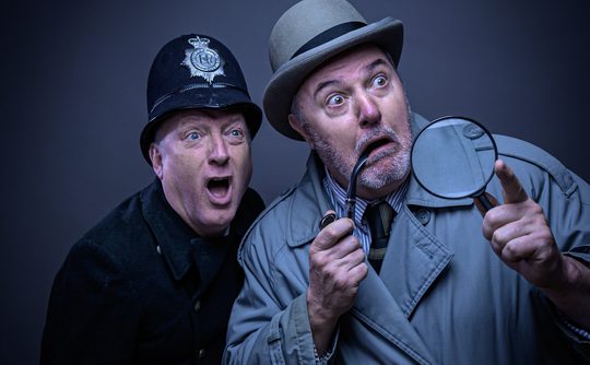Two men dressed as a detective and a policeman appear surprised. The detective wears a trench coat, holding a magnifying glass and pipe. Dramatic expressions.