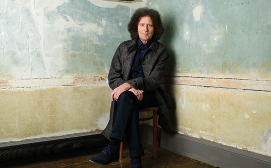 A man in a grey coat and blue shoes sits on a chair against a textured, faded wall. The wooden floor adds a rustic feel to the calm setting.