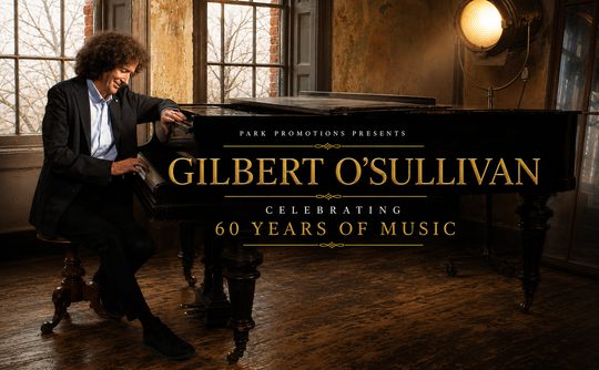 A joyful man plays a grand piano in an elegant, rustic room. Text on the piano reads "Gilbert O'Sullivan Celebrating 60 Years of Music."