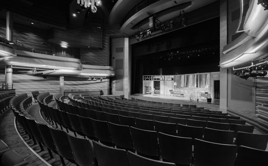 A black and white image of a proscenium arch theatre. On stage is the set of a living room.