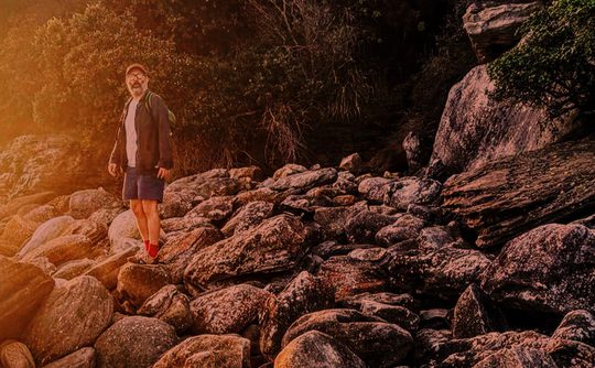 A man stands on a rocky shoreline at sunset, wearing a jacket, shorts, and red socks. The warm light casts a serene, adventurous mood.