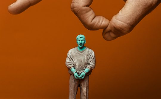 A hand hovers above a small, turquoise-tinged human figure wearing casual clothes against an orange background, creating a sense of control or manipulation.