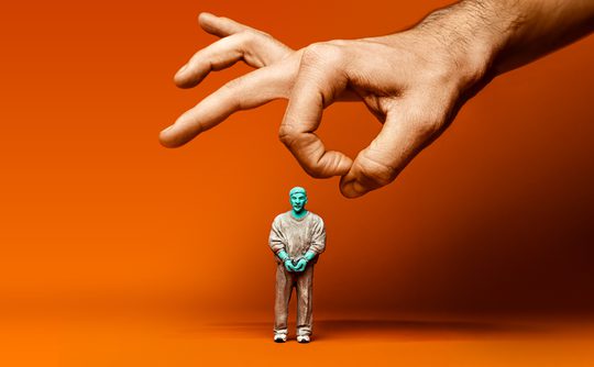A hand hovers above a small, turquoise-tinged human figure wearing casual clothes against an orange background, creating a sense of control or manipulation.