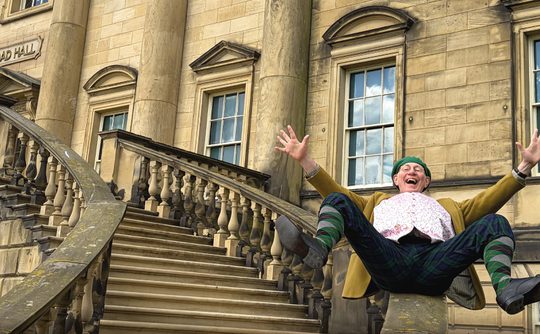 A joyful person in colorful attire sits playfully on a stone railing of an ornate staircase in front of Toad Hall, exuding excitement and energy.