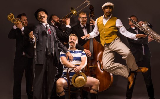 A lively jazz band with playful energy. One musician jumps mid-air, others play brass instruments. A seated man in a striped outfit holds a straw hat.