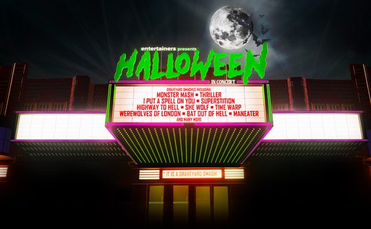 A concert marquee at night reads "Entertainers Presents Halloween In Concert" with a glowing green font. A full moon shines brightly above. Spooky ambiance.