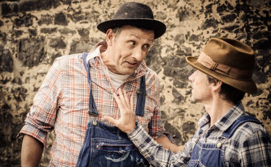 Two men in plaid shirts and denim overalls, wearing hats, engage in an animated discussion. One gestures with his hand on the other's chest. Stone wall backdrop.