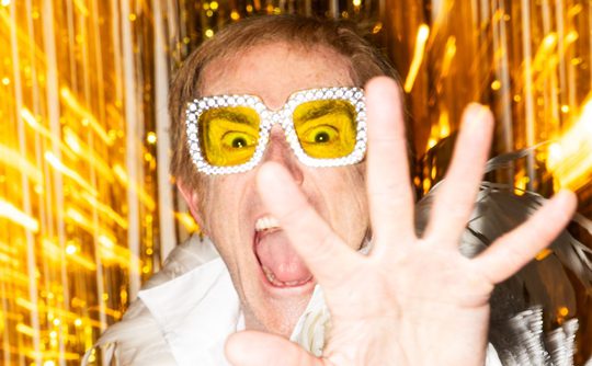 A person wearing sparkly yellow glasses and a white outfit excitedly reaches toward the camera. Bright gold streaks radiate in the background.