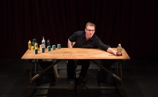 Man leaning over a wooden table on stage, surrounded by various cleaning products and props. He appears focused, under dramatic lighting, with a dark curtain in the background.