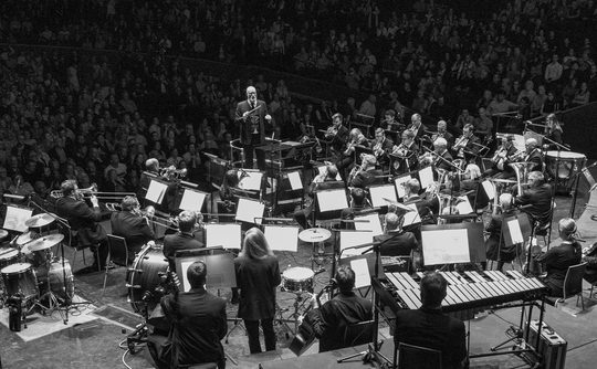 Black and white image of an orchestra performing on stage, with musicians intently playing various instruments. A conductor stands in front, leading. Audience fills the hall.