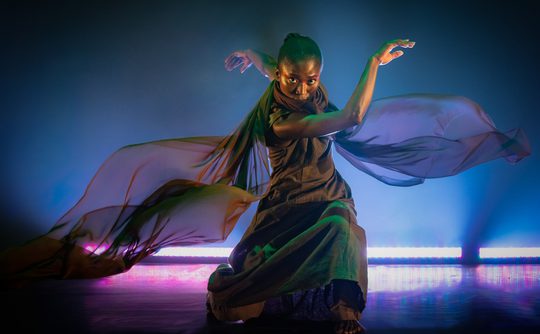 A dancer poses expressively with flowing fabric, bathed in blue and purple lighting. The scene conveys grace, movement, and intensity.