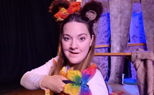 A woman dressed in a bear costume kneels on stage, holding a colourful rainbow butterfly. The background features tree-like props and scattered autumn leaves, creating a whimsical, theatrical setting.