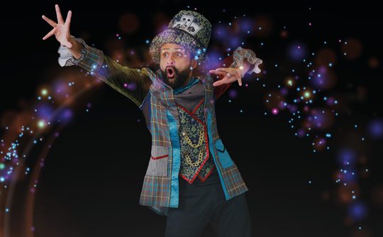 A man in a colourful, eclectic outfit and whimsical hat appears to cast a spell with a dramatic pose, surrounded by sparkling, magical lights.
