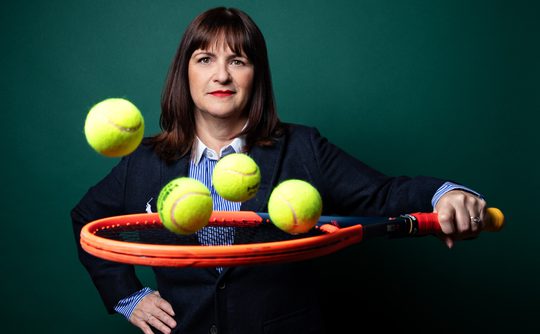 A woman in a blazer holds a tennis racket, with five tennis balls hovering mid-air. The background is a deep green, and her expression is confident.