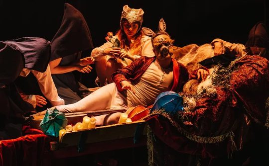 A dimly lit theatrical scene shows actors in medieval costumes. One actor reclines, surrounded by others wearing hoods and masks, creating a mysterious, dramatic atmosphere.