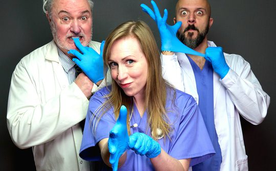 Three playful doctors wearing blue gloves and scrubs pose humorously. Two in lab coats act dramatically, while the woman in the center smiles confidently.