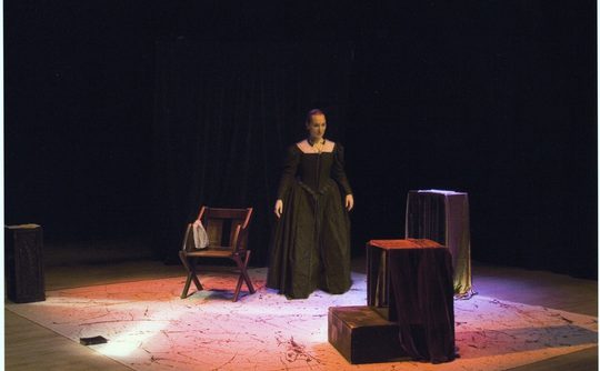 A woman in period costume stands on a dimly lit stage with a wooden chair and draped boxes. The atmosphere is dramatic and focused.