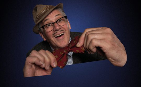 Older man with glasses and a moustache, smiling widely, adjusts a red bow tie. He wears a vintage hat and suit against a dark blue background.