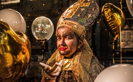 Drag King, Father JC, in ornate, glittery attire and dramatic makeup poses with balloons, exuding an extravagant, festive, and theatrical vibe.